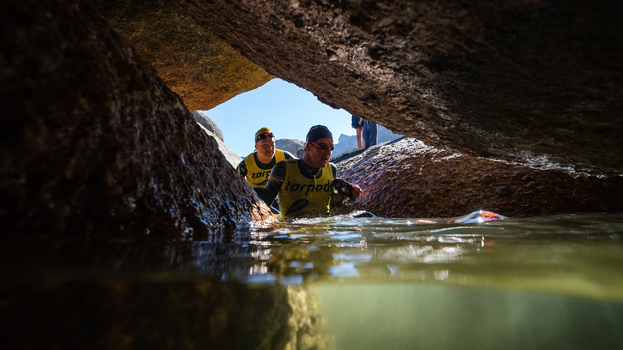 Two athletes wading through a narrow rock tunnel, seen from water level