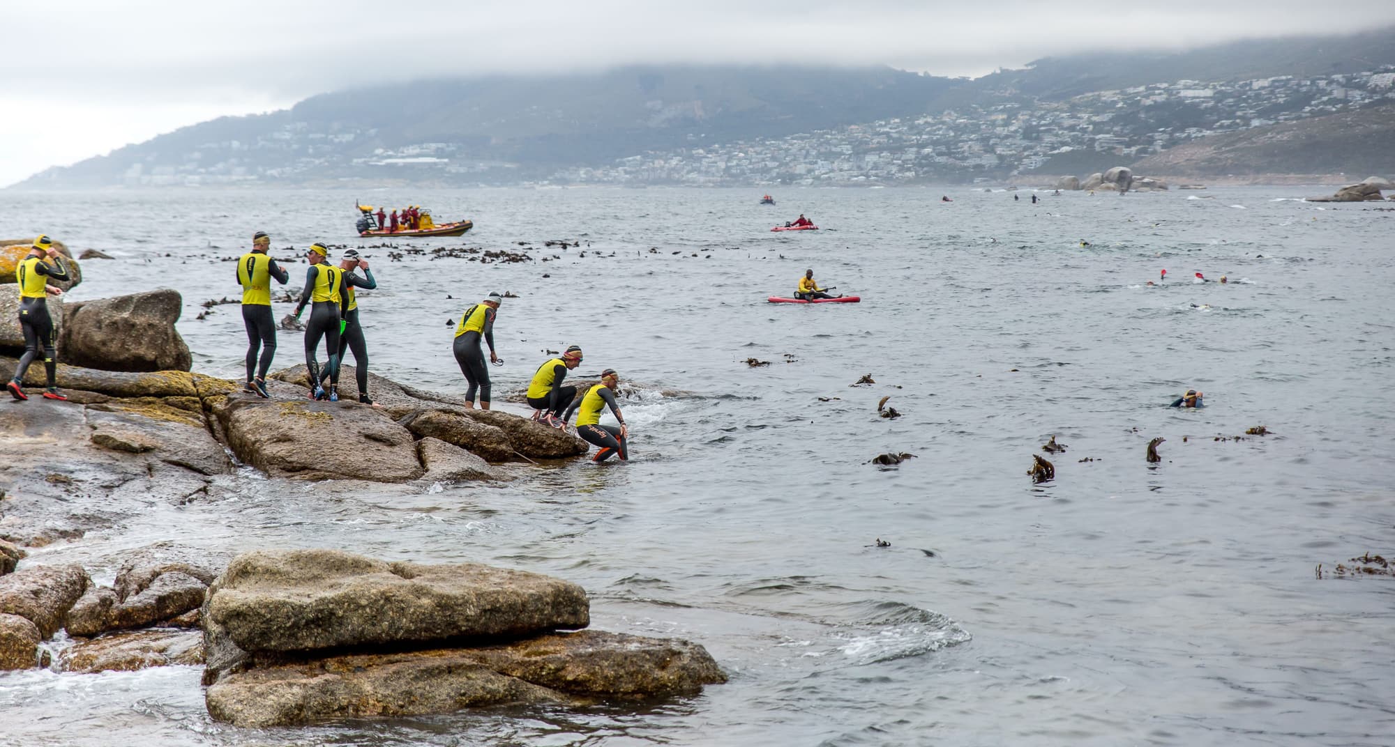 Athletes climbing over coastal rocks into the sea, with safety boats and Camps Bay behind